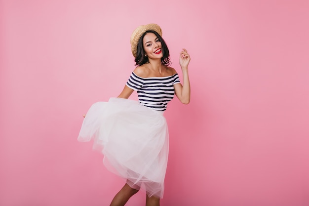 Shapely brunette lady in hat dancing with pleasure. Ecstatic female model in lush skirt and striped t-shirt having fun.