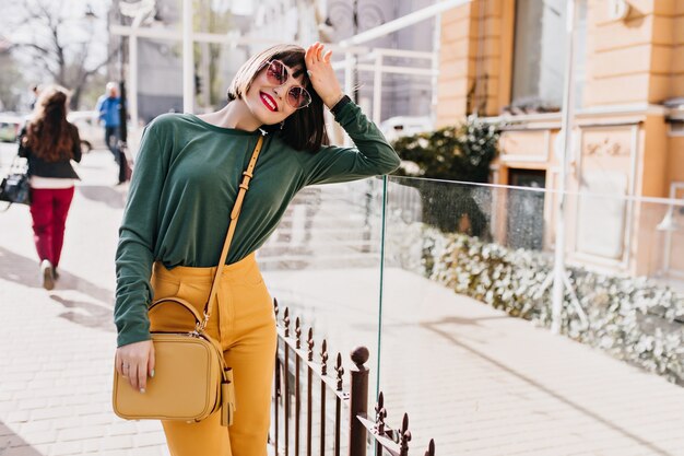 Shapely brunette woman with bright makeup standing on the street. Outdoor portrait of happy girl with trendy hairstyle posing in spring town.