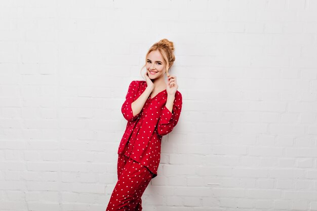 Shapely caucasian girl in red sleepwear playing with her hair. Laughing lady in cozy night-suit expressing true positive emotions.