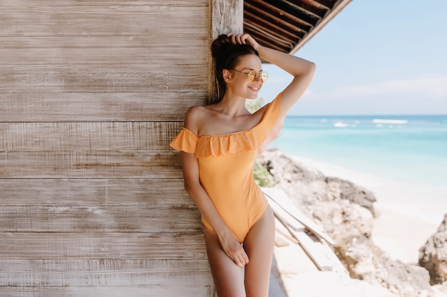 インスピレーションを得た顔でビーチでポーズをとるオレンジ色の服装の形の良い陽気な女の子。ビーチの木造住宅の横に立っているサングラスをかけた壮大な若い女性の屋外写真。