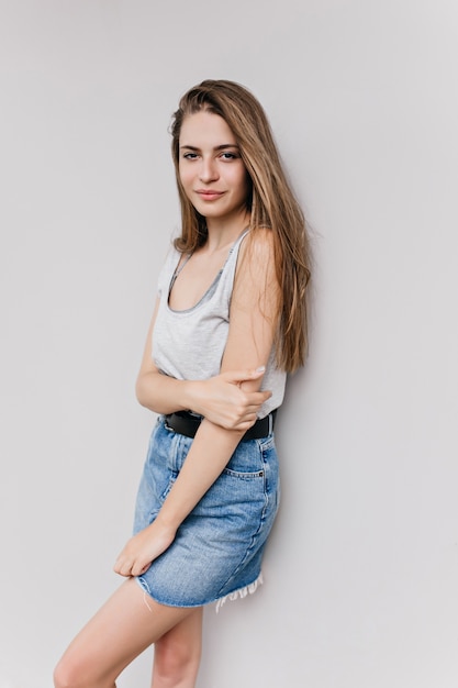 Shapely dark-haired woman standing near light wall and posing with interested face expression. Indoor shot of graceful caucasian lady wears denim skirt.