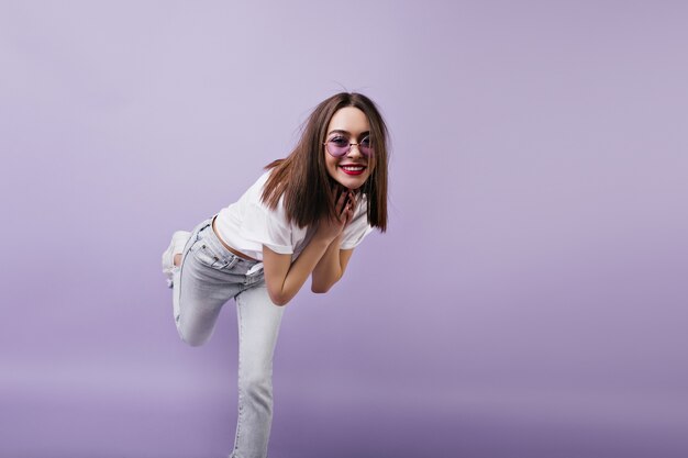 Shapely european woman in white jeans standing on one leg and fooling around. Indoor portrait of dancing female model in sunglasses.