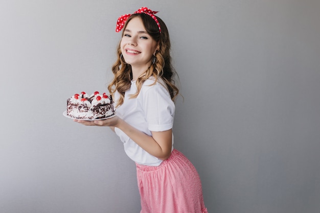 甘いパイを保持している赤いリボンを持つ形の良い女性モデル。バースデーケーキを持っている陽気な巻き毛の女性の屋内ショット。
