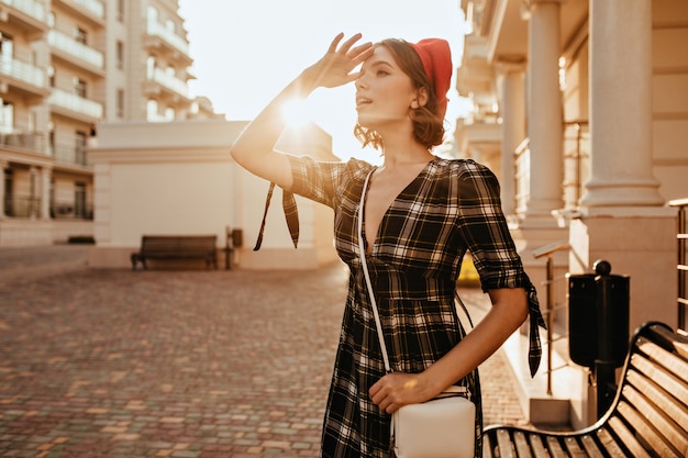 Shapely girl in elegant gray dress looking at distance. Outdoor portrait of lovable short-haired lady in red beret.