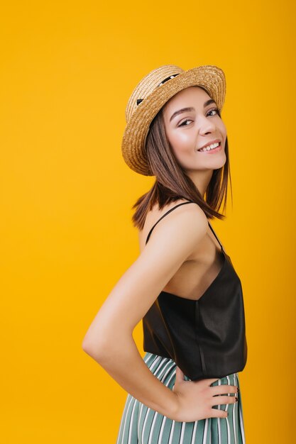 Shapely girl in elegant tank-top smiling. Indoor photo of female brunette model in straw hat.