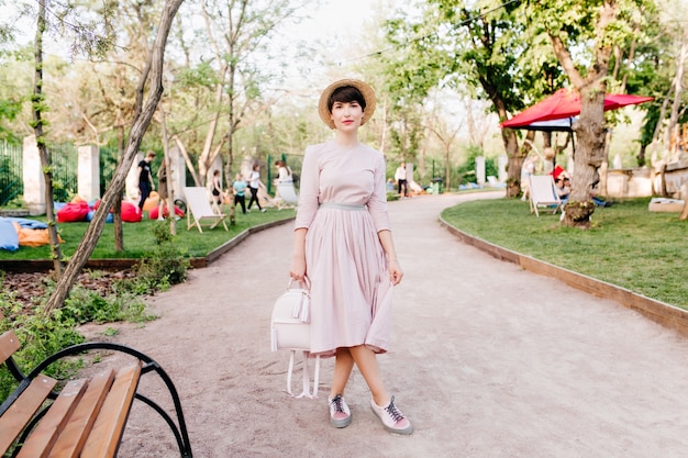 Shapely girl in trendy hat standing with legs crossed near the wooden bench in park