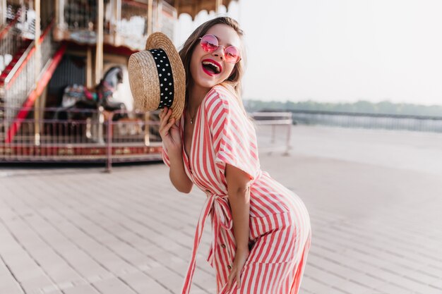 Shapely gorgeous lady in pink sunglasses expressing happiness in amusement park. Outdoor photo of laughing amazing girl standing near carousel.