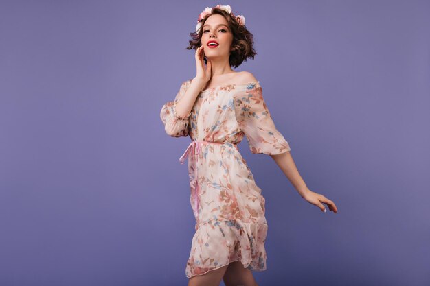 Shapely interested girl in summer dress standing on violet background Carefree caucasian young woman wears flowers in hair