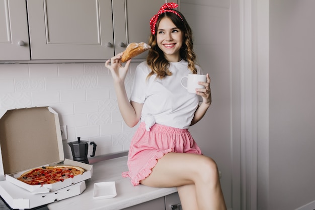 クロワッサンを食べるピンクのショートパンツで格好良い楽しい女の子。ピザとお茶を飲む巻き毛の髪型を持つ気さくな女性モデル。