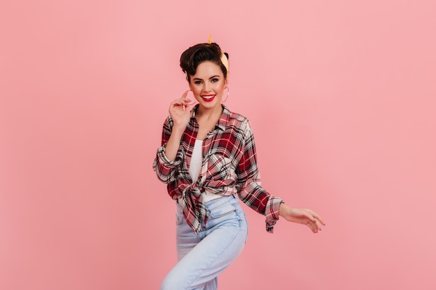 Shapely lady in jeans dancing on pink background. Studio shot of inspired pinup girl in checkered shirt looking at camera.
