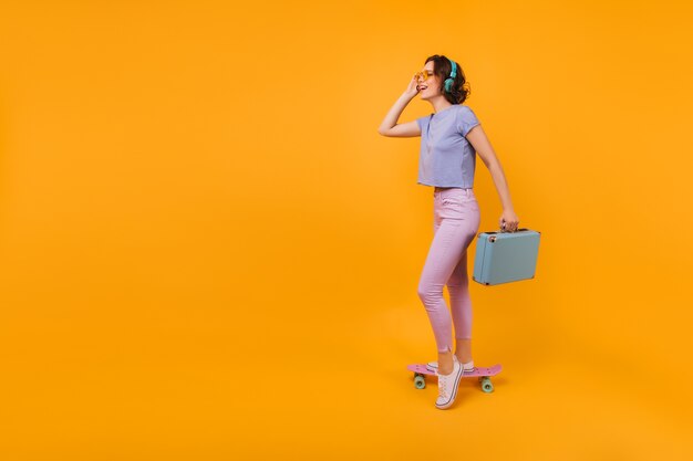 Shapely lady in white gumshoes posing with blue suitcase. Photo of good-humoured short-haired woman standing on longboard.