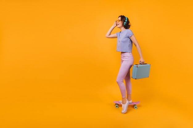 파란색 가방으로 포즈를 취하는 흰색 덧신에 매끈한 아가씨. longboard에 서있는 좋은 기분이 짧은 머리 여자의 사진.