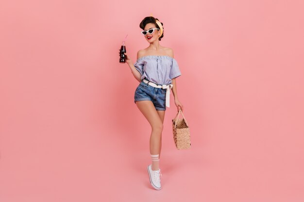 Shapely pinup girl holding bottle of soda and smiling. Stunning european woman with summer bag posing on pink background.