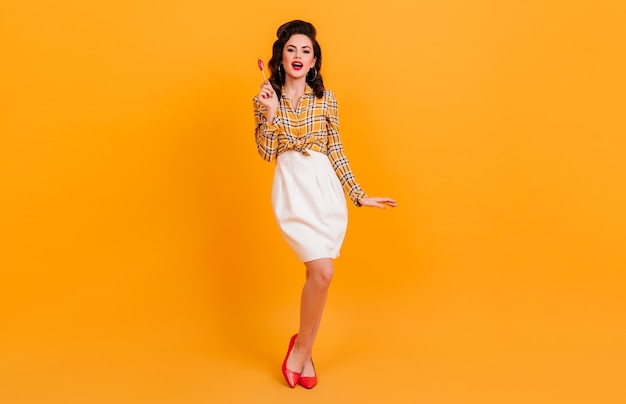 Shapely pretty woman in white skirt posing on yellow background. Studio shot of brunette pinup girl holding candy.