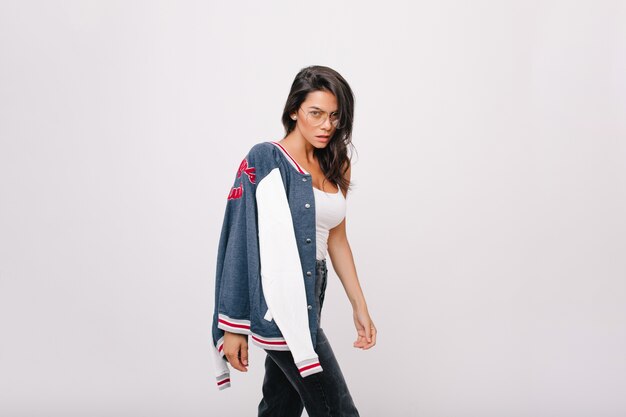 Shapely tanned girl in stylish glasses looking with interest while posing. Wonderful brunette european lady with sport bomber standing in front of white wall.