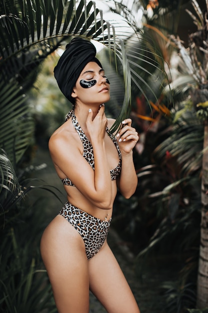 Shapely tanned woman in swimsuit touching neck on nature background. Outdoor shot of pretty woman in turban using eye patches.