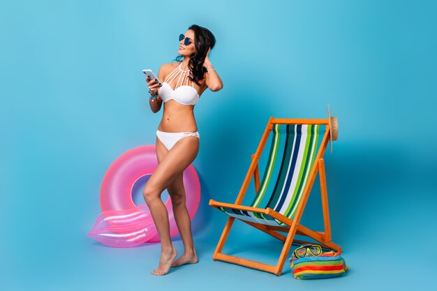 Shapely woman with smartphone standing beside deck chair