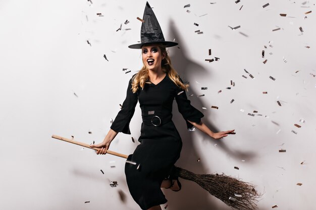 Shapely young witch in black attire sitting on broom. Indoor shot of cute wizard wears hat and long dress at halloween party.