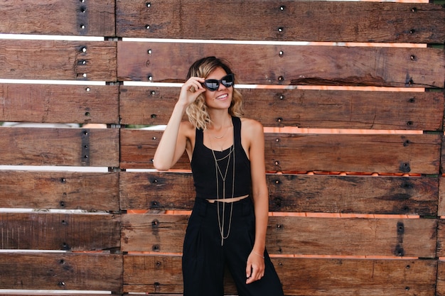 Shapely young woman in black pants having fun during photoshoot. pleasant curly female model wears sunglasses and stylish tank-top.