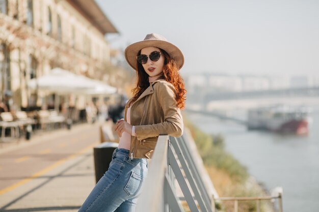 Shapely young woman in hat standing near sea and looking to camera