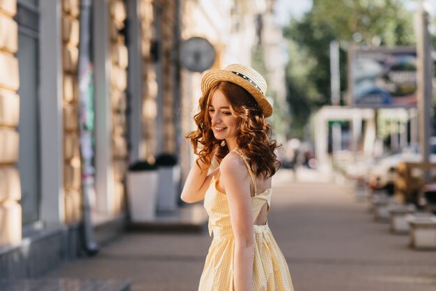 도시에서 기쁨과 함께 포즈 노란색 드레스에 매끈한 젊은 여자. 산책하는 동안 사진 촬영을 즐기는 모자에 멋진 여자의 야외 사진.