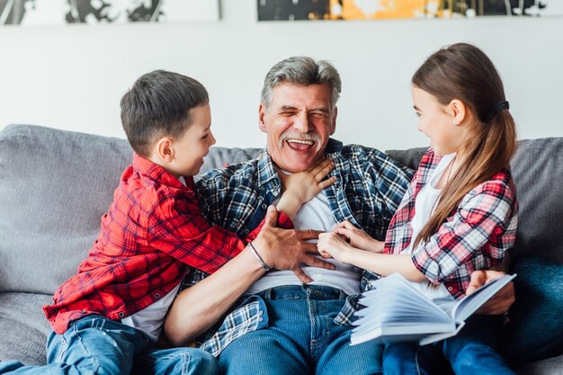 Поделись своей мудростью. Позитивный пожилой мужчина читает книгу для своих внуков, отдыхая дома.