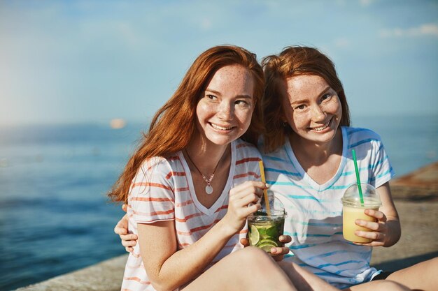 최고의 순간을 공유하는 빨간 머리와 주근깨를 가진 두 명의 멋진 소녀가 해변 근처에 앉아 칵테일을 마시고 껴안고 편안하고 행복한 느낌으로 함께 시간을 보내는 것을 즐깁니다.