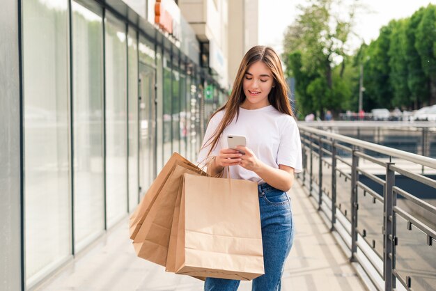 良いたよりを友達と共有する。屋外に立っている間買い物袋と携帯電話を保持している美しい若い笑顔の女性のクローズアップ