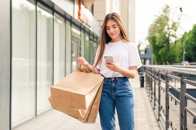 Condividendo buone notizie con un amico. primo piano di bella giovane donna sorridente che tiene i sacchetti della spesa e il telefono cellulare mentre levandosi in piedi all'aperto