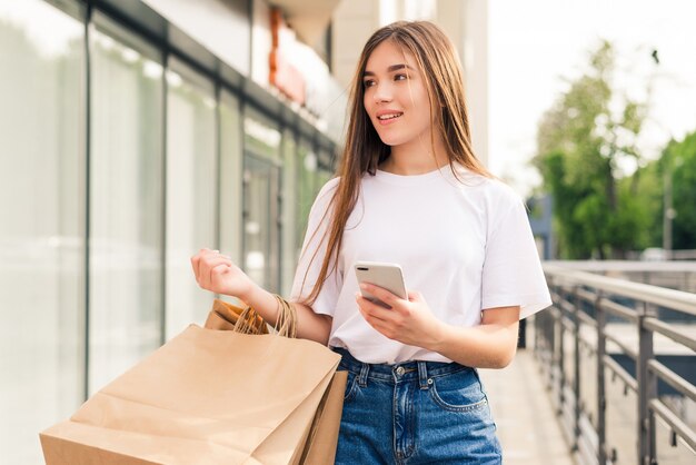 良いたよりを友達と共有する。屋外に立っている間買い物袋と携帯電話を保持している美しい若い笑顔の女性のクローズアップ