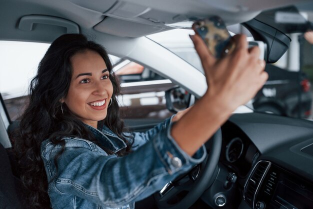 幸せを共有します。自動車サロンで彼女のブランドの新しい高価な車を試す黒い髪のかわいい女の子