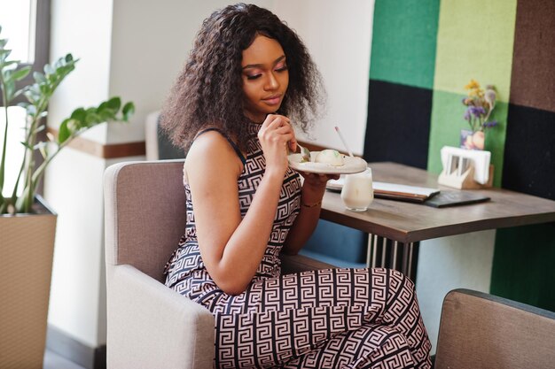 Sharming elegant young african american woman with long curly hair wearing jumpsuit posing at cafe indoor with panna cotta dessert