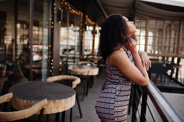 Sharming elegant young african american woman with long curly hair wearing jumpsuit posing at cafe terrace outdoor against christmas decoration New Year eve