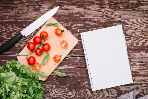 Sharp knife; basil; cherry tomatoes and lettuce with spiral notepad on wooden surface