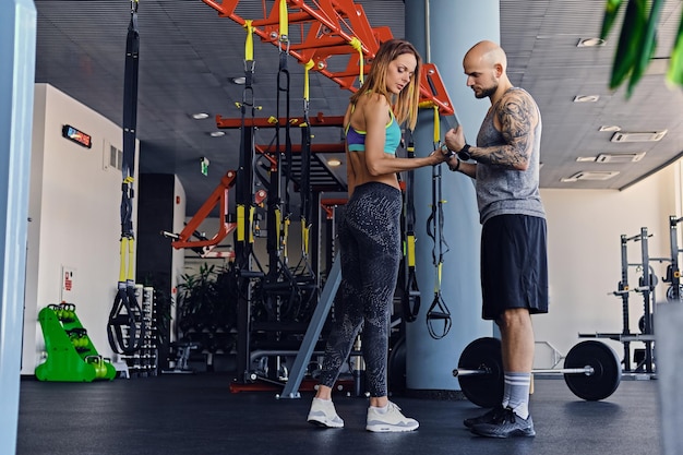 Shaved head athletic male and slim brunette female exercising with trx straps in a gym club.