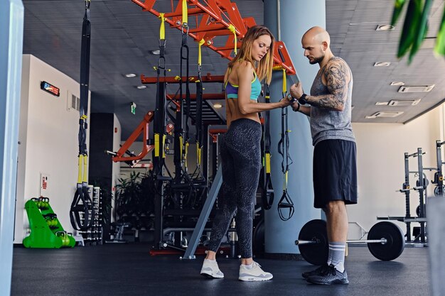 Shaved head athletic male and slim brunette female exercising with trx straps in a gym club.