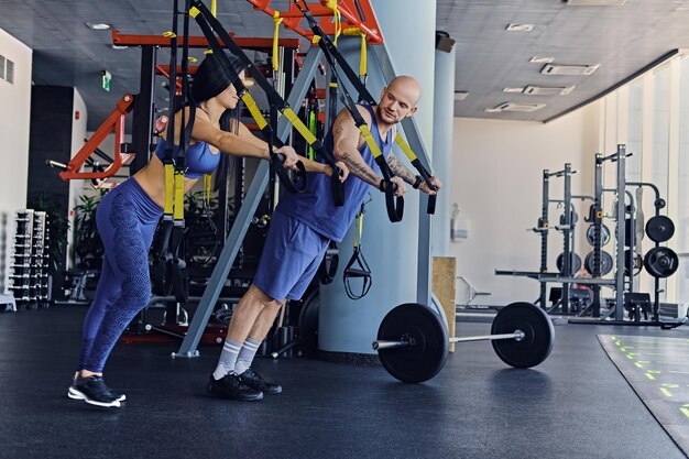 Shaved head athletic male and slim brunette female exercising with trx straps in a gym club.