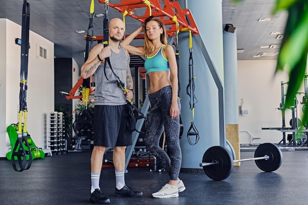 Shaved head athletic male and slim brunette female posing near trx straps stands in a gym club.