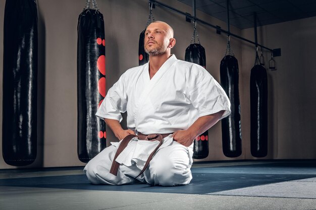 Shaved head karate fighter sits on the tatami in a gym.