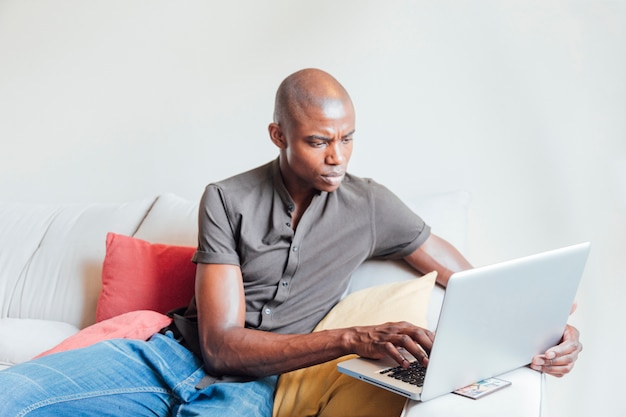 Shaved young african man sitting on sofa using laptop