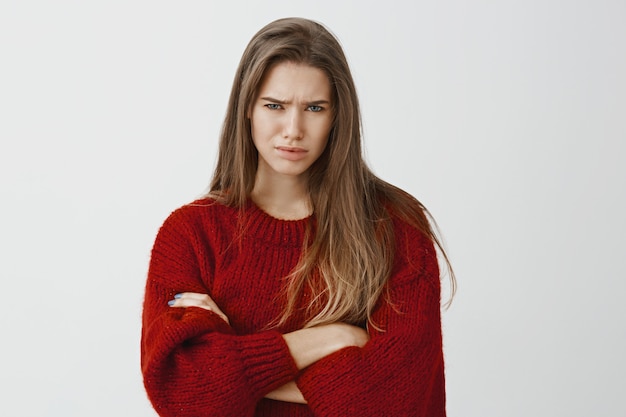 She do not buys on such clumsy pick-up lines. Doubful displeased european female student in red loose sweater, crossing hands and frowning, expressing disbelief and frustration over gray wall