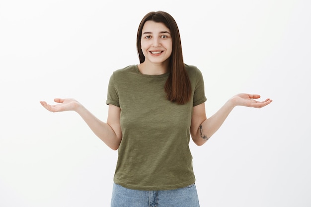 She has no idea. Portrait of cute and friendly attractive young 20s caucasian girl making shoudler shrug as spread hands sideways in clueless pose, smiling silly being uanware over gray wall