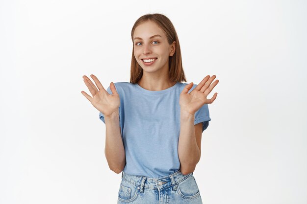 She is not involved. Smiling blond girl raising empty hands up and looking careless, nothing to say, being unaware and clueless, standing in t-shirt against white background