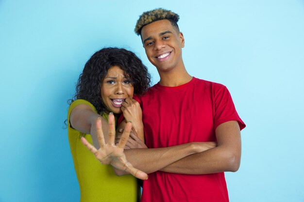 She's scared, he's laughting. Young emotional african-american beautiful man and woman in colorful clothes on blue background.