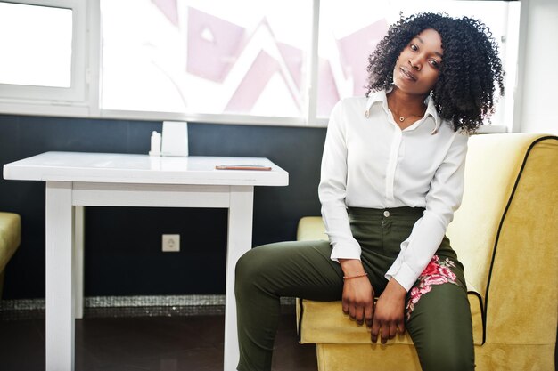 Sheerful business african american lady with afro hair wear white blouse and green pants posed in cafe