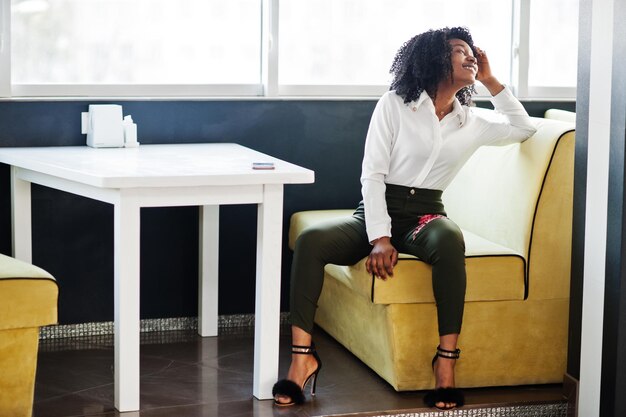 Sheerful business african american lady with afro hair wear white blouse and green pants posed in cafe
