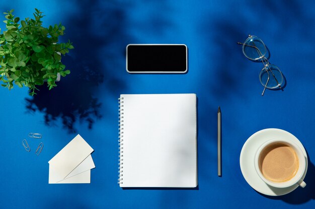 屋内の青いテーブルにシート、コーヒー、作業工具。ホームオフィスの創造的で居心地の良い職場、表面に植物の影が付いたインスピレーションあふれるモックアップ。リモートオフィス、フリーランス、雰囲気のコンセプト。