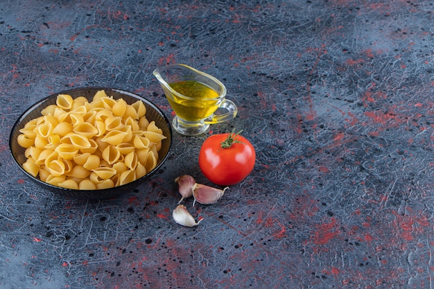 Shell uncooked pasta in a glass bowl with fresh red tomato and garlic .