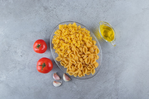 신선한 빨간 토마토와 마늘이 든 유리 그릇에 생으로 건조된 Ditali Rigati와 함께 조리하지 않은 파스타를 껍질을 벗기십시오.