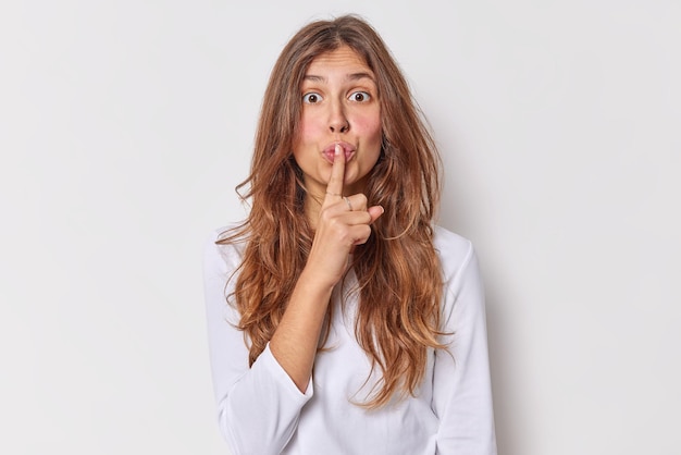 Shh its my secret. Surprised long haired woman tells be silent keeps index finger pressed over lips makes shut up gesture dressed casually isolated over white background gossips and spreads rumors